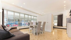 Una sala de estar moderna con sofá, mesa de comedor, sillas y ventanas amplias con vistas a la ciudad.