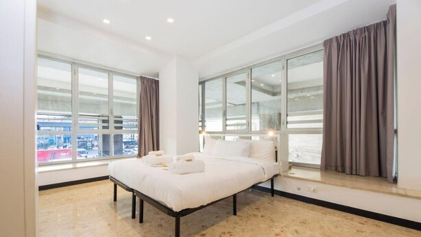 Una habitación de hotel moderna con una cama grande, dos ventanas con cortinas y vistas a la ciudad.