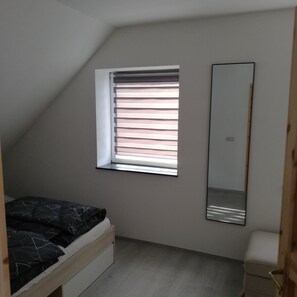 Een slaapkamer met een schuine plafond, een raam met jaloezieën, een bed met hoofdeinde, een nachtkastje en een spiegel aan de muur.