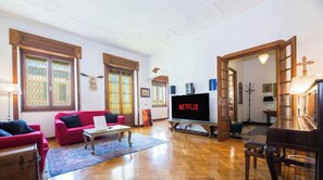 Ein Wohnzimmer mit einem roten Sofa, einem Couchtisch, einem Fernseher, der 'NETFLIX' zeigt, und einem Klavier.