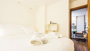 Ein Hotelzimmer mit einem ordentlich bezogenen Bett, einer kleinen Ablage mit einer Lampe und Blick auf den Flur.