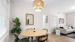 Ein modernes Wohnzimmer mit einem runden Holztisch, zwei Stühlen, einer Couch, einem großen Spiegel und einer hängenden Leuchte.