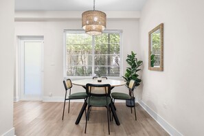 A dining area with a round table, four chairs, a hanging light fixture, a large window with blinds, and a mirror on the wall. - Chic Sobe Retreat 2-bedroom 2 full bath apartment walk to Lincoln Rd & Bay (Miami Beach)