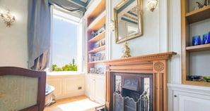 Ein Zimmer mit Kamin, einem fensterbehangenen Fenster, einem Bücherregal und einem Wandspiegel.