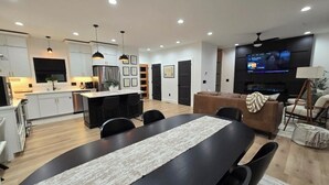 Cozinha moderna com armários brancos, sala de jantar com mesa preta e sala de estar com sofá marrom e televisão.
