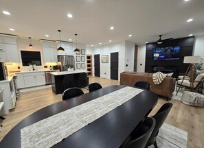 Eine moderne Küche mit weißen Schränken, ein Essbereich mit einem schwarzen Tisch und ein Wohnzimmer mit einem braunen Sofa und einem Fernseher.