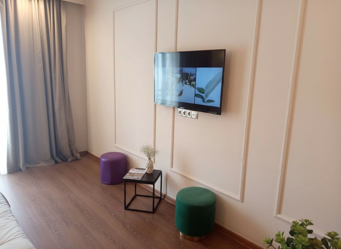 A modern room with a mounted TV, a small table with a vase, a green ottoman, and a wooden floor.