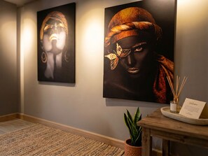 A hallway with two framed artworks on the wall, a small wooden table with a plant and a bottle, and a woven rug on the floor. - Riverside Central Apartments (T'bilisi)