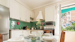 A kitchen with white cabinets, a green tiled backsplash, a dining table set with plates and glasses, and a view of buildings through a window. - Beach 7M| 1BR| Wifi| Nervi Park By Tilo (Genova)