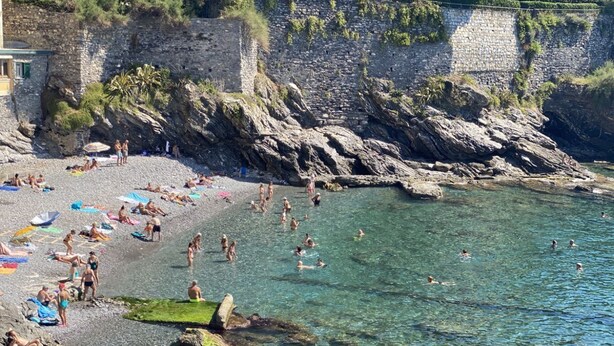 Uma praia rochosa com águas cristalinas, pessoas tomando sol e nadando, e um muro de pedra ao fundo.