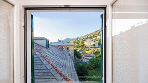 Visão através de uma janela de um edifício com telhado de azulejos, cerca verde e um morro com construções.