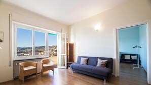 Quarto com sofá, duas cadeiras de vime, vista pela janela grande e porta que dá para outro cômodo.