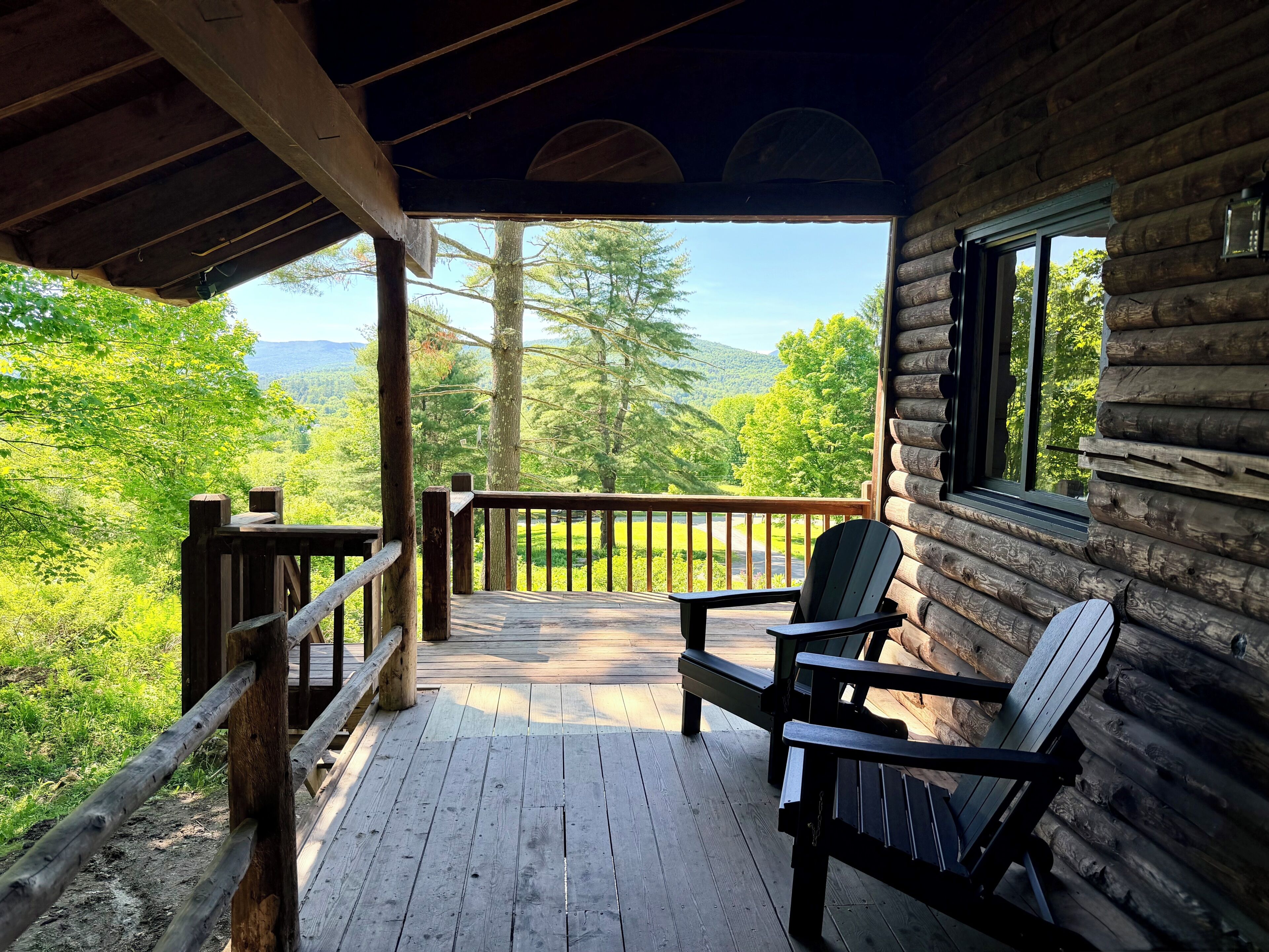 En träveranda med två Adirondack-stolar, ett träräcke och ett fönster med utsikt över ett skogslandskap.