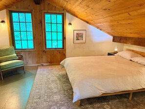 Ein Schlafzimmer mit Holzboden, einem Bett mit gestreifter Bettdecke, einem Sessel und zwei Fenstern mit Blick.