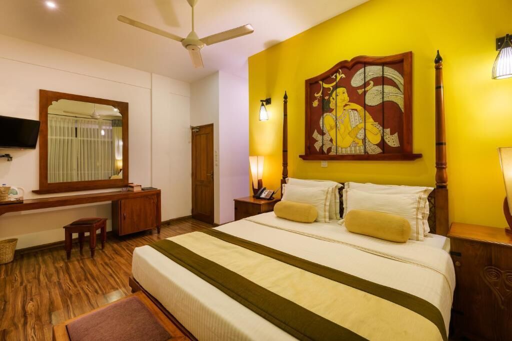 Une chambre d’hôtel équipée d’un lit, d’un bureau, d’une chaise, d’une télévision, d’un ventilateur de plafond et d’une œuvre d’art accrochée au mur.