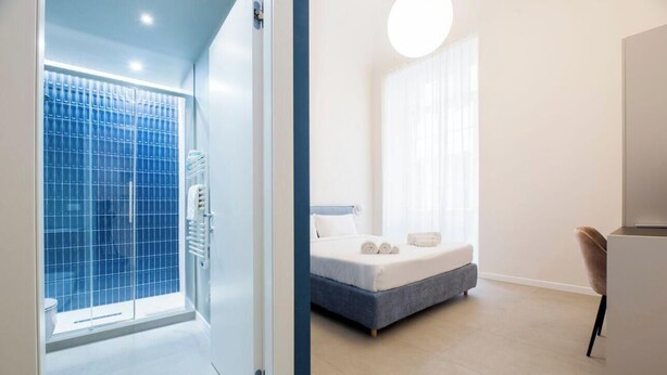 A modern bedroom with a bed, a chair, and a bathroom with a shower. - Aquarium 1'| 3BR| A/C|Parking| City Center By TILO (Genova)