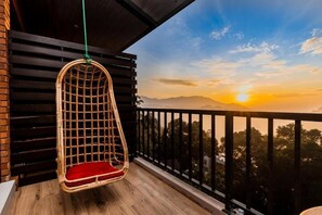 Una silla colgante de mimbre con un cojín rojo en un balcón de madera con vistas a una puesta de sol y montañas.
