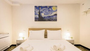 A hotel room with a bed, two bedside tables with lamps, a painting on the wall, and a white air conditioning unit. - AQUARIUM 1'|1BR|A/C| WIFI|BY TILO (Genoa)