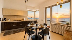 Een moderne keuken met een eetgedeelte, grote ramen met uitzicht op een zonsondergang en een balkon met uitzicht op zee.
