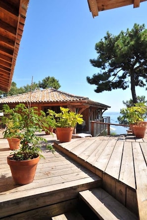 Une terrasse en bois agrémentée de plantes en pot, une structure surmontée d’un toit en tuiles et une vue sur la mer.
