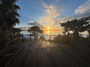Une terrasse en bois offrant une vue sur le coucher de soleil sur l’eau, entourée d’arbres et d’une clôture.