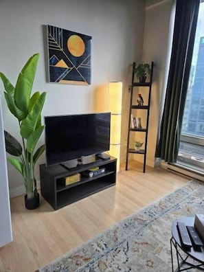 Ein modernes Wohnzimmer mit einem Flachbildfernseher, einer großen Topfpflanze, einem Bücherregal und Blick auf die Stadt durch ein Fenster.