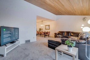 A modern living room with a flat-screen TV, leather sofa, dining area, and a ceiling with wooden beams.