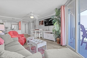 Ein Wohnzimmer mit einer Couch, Sesseln, einem Couchtisch, einem Fernseher, einer Pflanze und einem Balkon mit einem Stuhl.