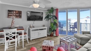 Een woonkamer met een televisie, witte meubels, een plafondventilator, een plant en een balkon met stoelen.