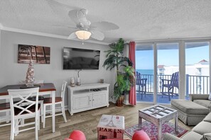 Ein Wohnzimmer mit Fernseher, weißen Möbeln, Deckenventilator, Pflanze und einem Balkon mit Sitzgelegenheiten.