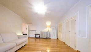 Ein geräumiges Zimmer mit Holzboden, einem weißen Sofa, einem Tisch und einer Lampe. Weiße Türen und ein Deckenventilator sind vorhanden.