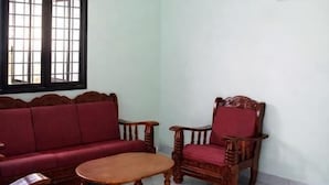 Una habitación con ventilador de techo, sofá rojo, una silla de madera, una mesita y una ventana con barrotes.