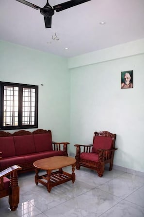 A room with a ceiling fan, a red sofa, a wooden chair, a small table, and a window with bars. - Bhagawan Guest House (Tiruvannamalai)