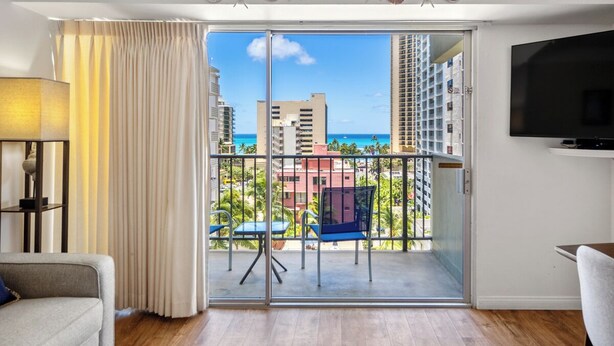 Ein modernes Wohnzimmer mit Balkon, einer Couch, einem Fernseher und Blick auf das Meer und Gebäude.