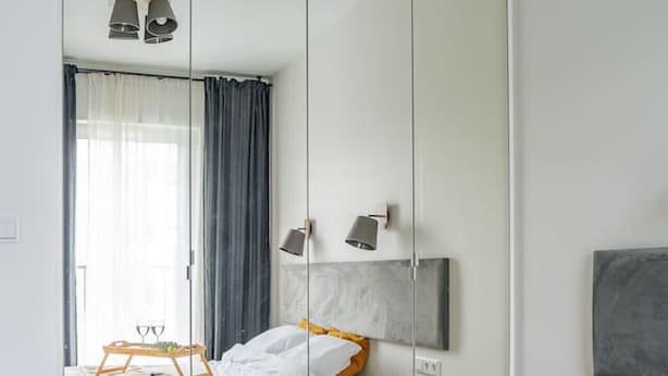 A hotel room with a large mirror, a bed with white linens, a bedside table with a lamp, and a tray with glasses. - Rakowicka Apt - Main Station, Old Town nearby - by Rentujemy (Kraków)