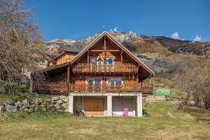 A wooden chalet with a balcony, a garage, and a mountainous backdrop.