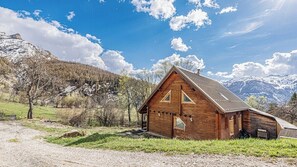 A wooden cabin with a sloped roof, surrounded by a gravel path, trees, and mountains.