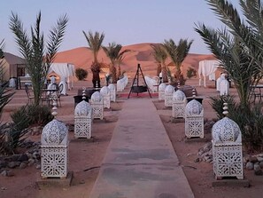 Un sentier désertique menant à un espace aménagé avec des tentes, des tables et des chaises, entouré de palmiers et illuminé de lanternes décoratives.