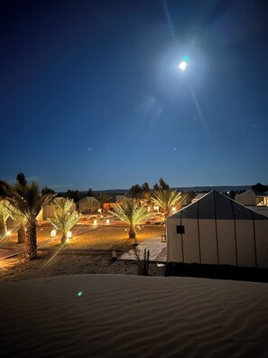 Un campement dans le désert, de nuit, avec des tentes, des palmiers et une lune brillante.