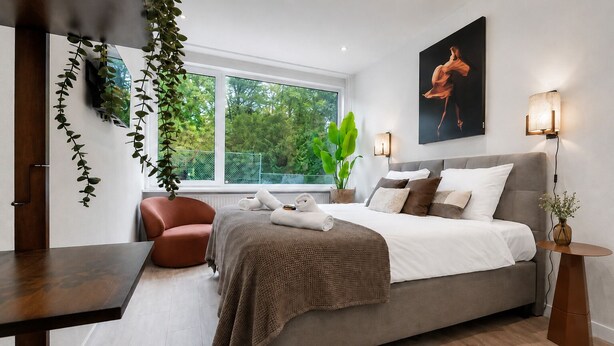 A modern bedroom with a large bed, a chair, a table, and a window with a view of greenery.