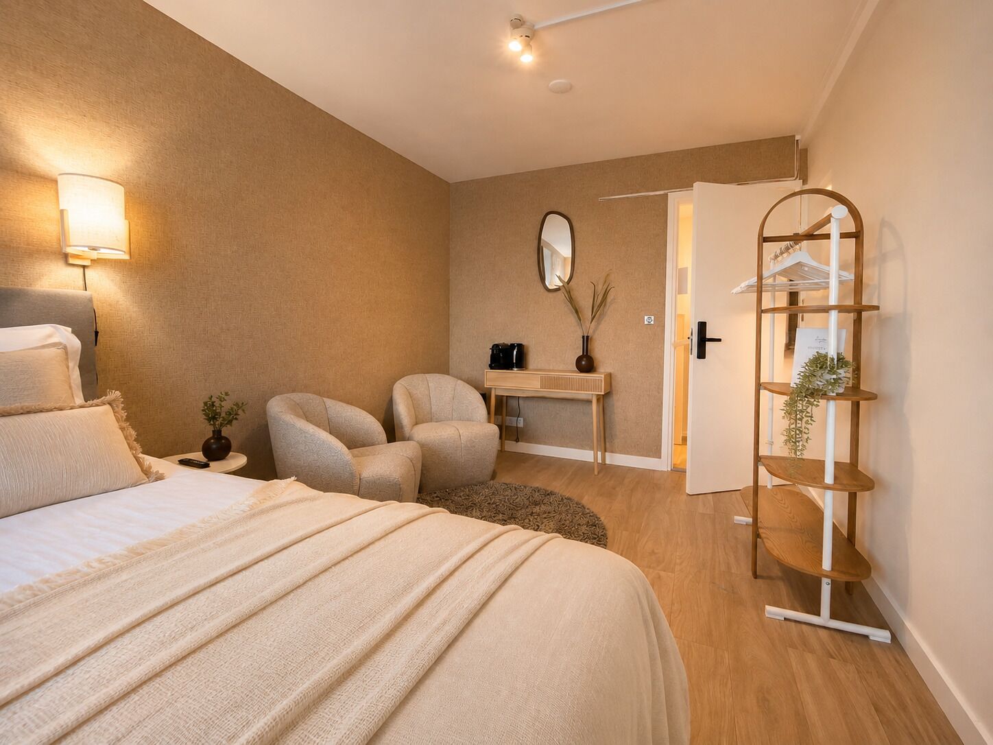 Un dormitorio con una cama, dos sillas, una mesita con un espejo y un estante de madera con plantas.