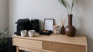 A wooden desk with a coffee maker, two cups, a coffee guide, a jar of nuts, a vase with reeds, and a potted plant.
