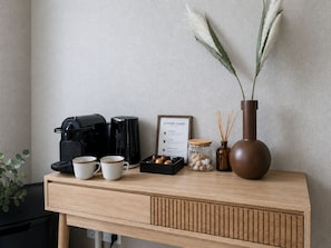 Et træbord med en kaffemaskine, to kopper, en kaffeguide, en krukke med nødder, en vase med rør og en potteplante.