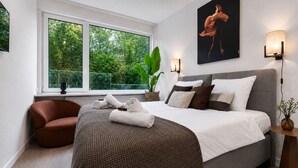 A modern bedroom with a large bed, a brown armchair, a window with a view of greenery, and a painting on the wall.