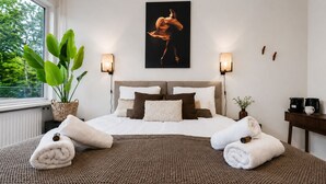 A bedroom with a bed, two bedside lamps, a potted plant, a coffee maker, and a painting on the wall.