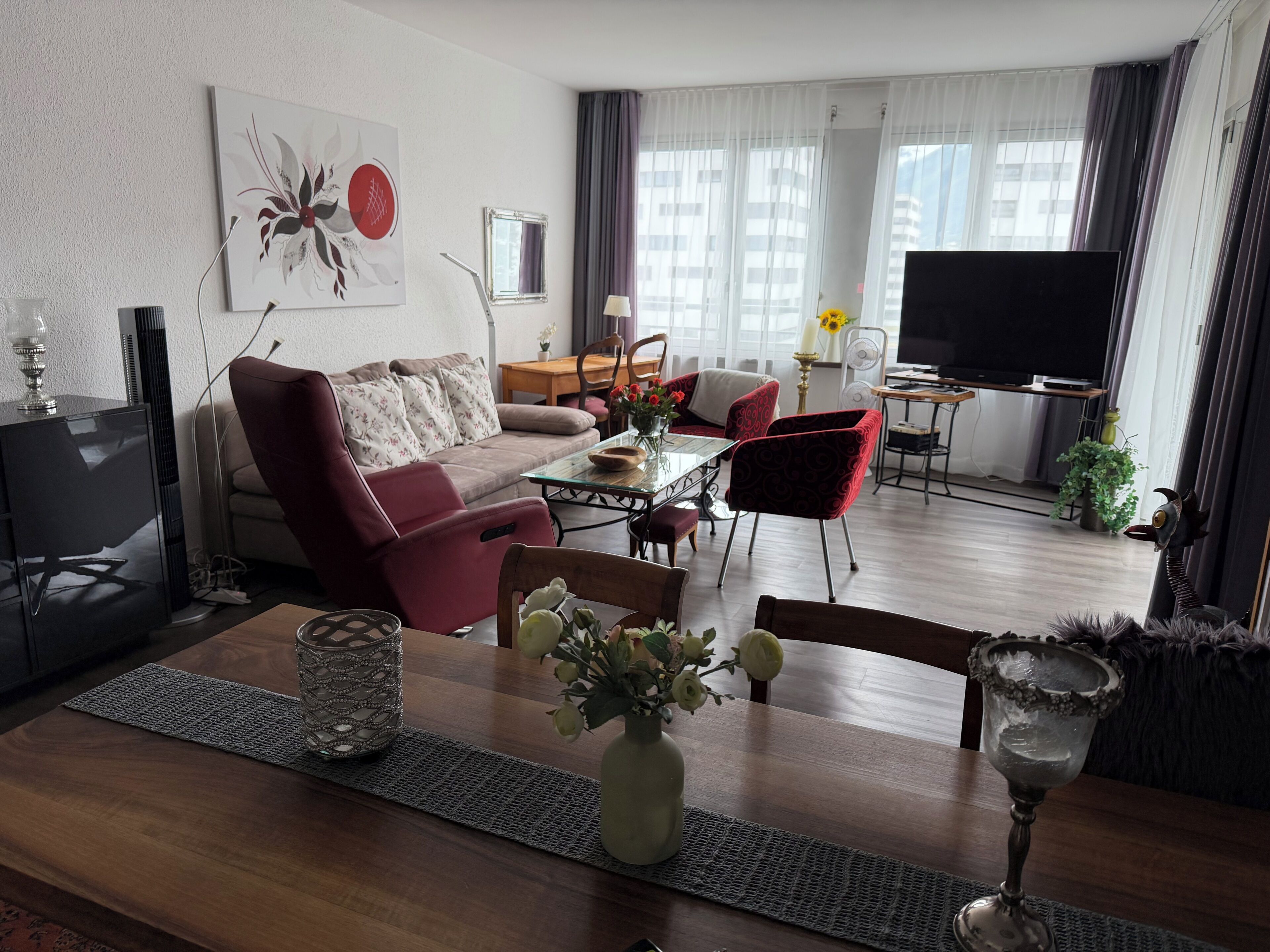 A modern living room with a sofa, chairs, a dining table, a TV, and a large window with curtains.