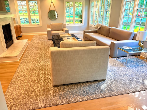 A modern living room with a fireplace, two sofas, a coffee table, and large windows with a view of greenery.