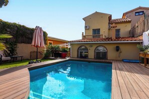 Ein Schwimmbad mit Holzterrasse, einer Terrassenfläche und einem Gebäude mit Ziegeldach.