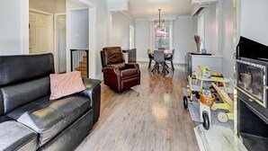 Un salon comprenant un canapé en cuir noir, un fauteuil en cuir marron, un coin repas avec une table et des chaises, ainsi qu’un meuble pour la télévision avec un chariot.