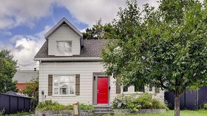 Une maison à deux étages avec une porte rouge, un petit jardin devant et un arbre.